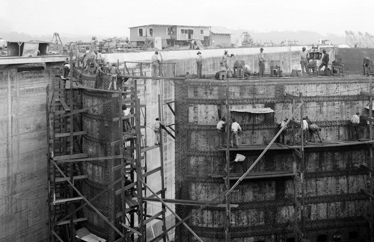 Black-and-white photo of a huge metal gate with tiny workers either posing or working from the wooden scaffolding.