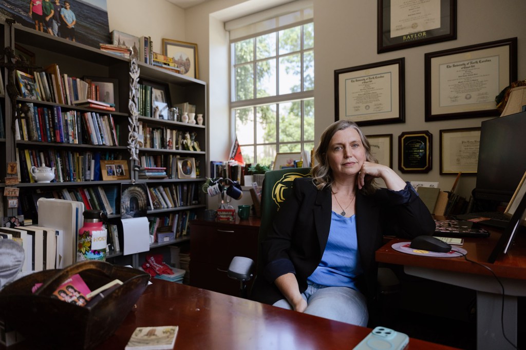 A woman sits in an office at a desk, with bookshelves, framed diplomas, and a window behind her.