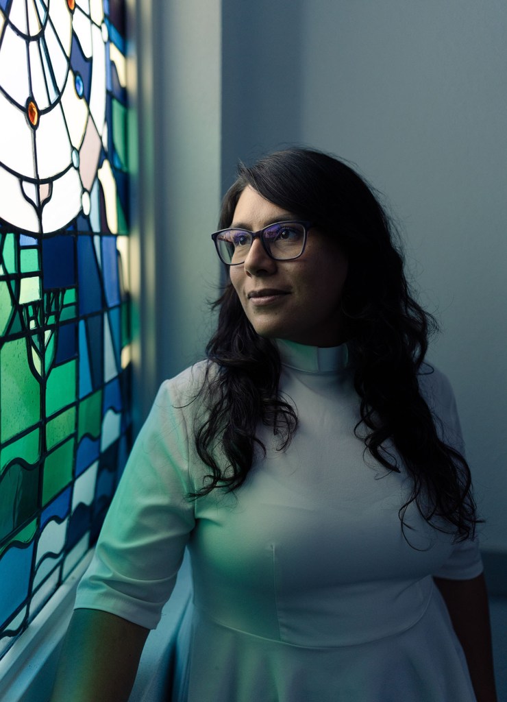 A woman wearing glasses and a white clerical collar stands beside a stained glass window, looking out.