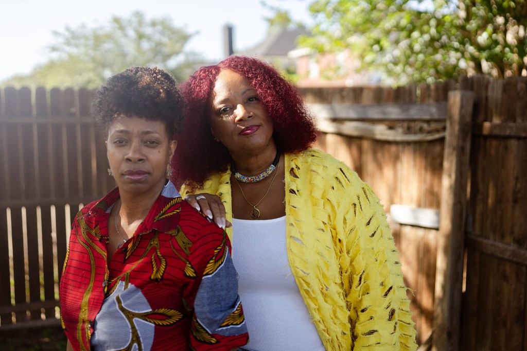 Two women stand close together outdoors in front of a wooden fence, one resting her hand on the other’s shoulder.