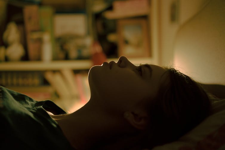 Teenage girl lying on her back in bed in near darkness with her eyes open.