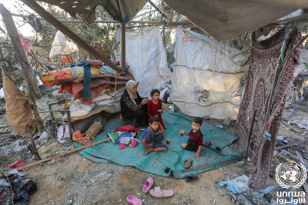 A Palestinian woman sits with her children in front of her house destroyed by the Israeli bombing that continue on Deir el-Balah, Gaza Strip. 7 August 2024. United Nations Relief and Works Agency.