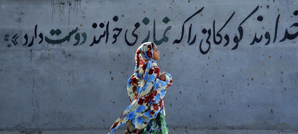 An Iranian girl in a colorful headscarf and dress runs past a wall with a Persian mural in the village of Bahl.